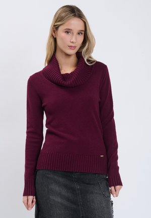 Jeune femme portant un pull à col roulé prune à manches longues et une jupe en jean foncée, debout devant un fond clair uni.