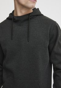 Felpa con cappuccio di un grigio scuro, realizzata in tessuto strutturato, con cappuccio regolabile e tasca frontale. Presenta una vestibilità rilassata e un design minimalista.