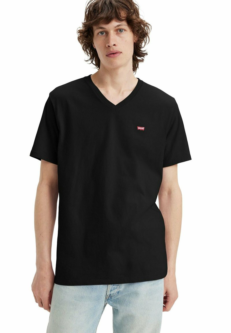 Junge Person mit welligem braunem Haar trägt ein schwarzes Levi’s T-Shirt mit V-Ausschnitt und hellblauen Jeans, steht vor weißem Hintergrund.