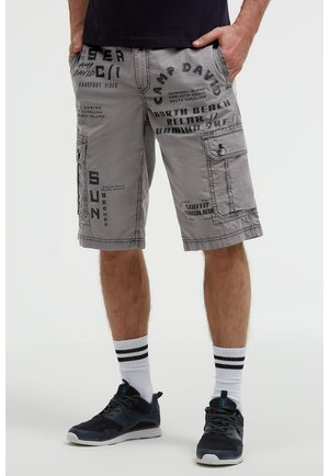Mann trägt graue Cargoshorts mit schwarzem Aufdruck, weiße Socken mit schwarzen Streifen und schwarze Sportschuhe, steht mit den Händen in den Taschen.