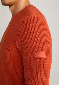 Maglione arancione lavorato a maglia con texture a costine, collo rotondo e una etichetta in pelle sulla manica sinistra.