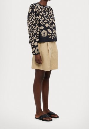 Personne portant un haut noir à manches longues avec des motifs floraux beiges, un short beige jusqu'aux genoux et des sandales noires à enfiler, debout sur un fond blanc.