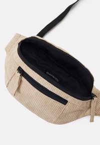 Marsupio in corduroy beige con cerniera nera e fodera interna. Presenta una superficie texturizzata e una tracolla nera regolabile.