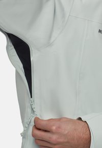 Veste imperméable gris clair avec fermeture éclair et doublure intérieure noire. Comprend un tissu texturé et un cordon de serrage réglable sur l'ourlet.