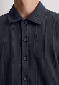 Donkerblauwe blouse met een gestructureerde, geribbelde stof en een platte kraag. Voorzien van zes afgeronde knopen langs de voorpaspel.