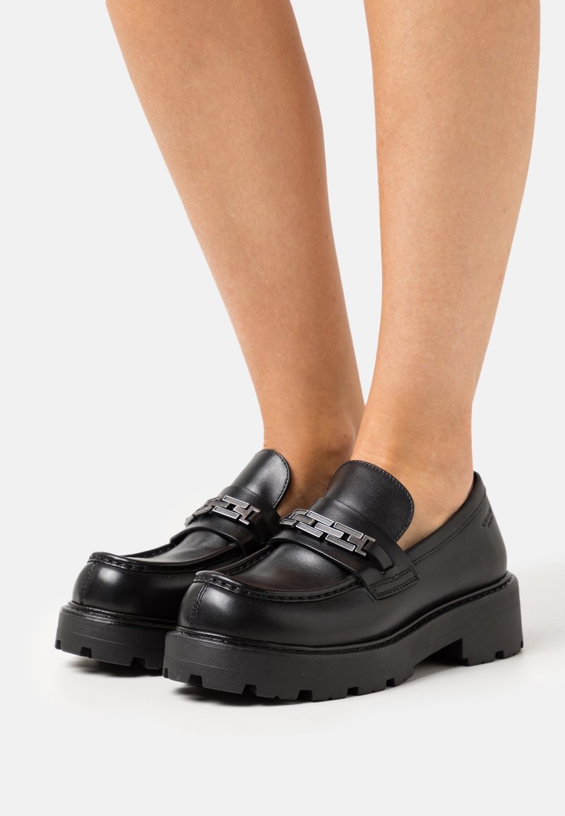 Mocassins noirs à semelle épaisse avec un détail de chaîne argentée, portés pieds nus sur un fond blanc uni.