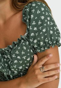 Grønt blomstermønsteret bluse med korte flæsekorte ærmer og snor til halsen. Glat tekstur med en subtil krølleeffekt.