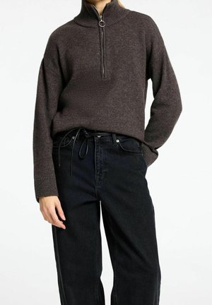 Personne portant un pull en tricot marron foncé à fermeture éclair, rentré dans un jean noir taille haute avec une ceinture en corde noire, sur fond blanc uni.