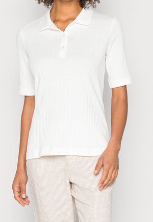Person trägt ein weißes kurzärmeliges, geripptes Poloshirt und helle beige gestreifte Hose und steht vor einem einfarbigen Hintergrund.