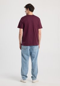 Man wearing a maroon t-shirt, light blue jeans, and white sneakers, standing facing away against a plain white background.