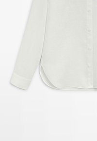 Camicia bianca in lino a maniche lunghe con polsino abbottonato e orlo arrotondato, con chiusura frontale a bottoni su sfondo semplice.