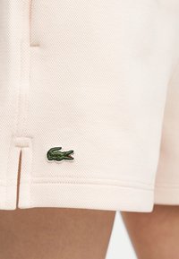 Short en tissu texturé rose clair avec un petit logo crocodile vert brodé près de l'ourlet sur la cuisse d'une personne.