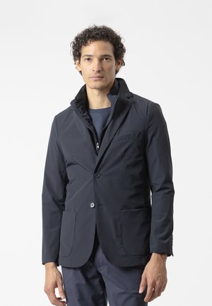 Hombre con cabello rizado que lleva una chaqueta negra con capas y una chaqueta con cremallera, de pie frente a un fondo claro y liso.