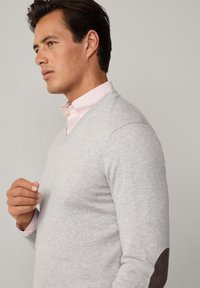 Grauer V-Ausschnitt-Pullover aus weichem Material, mit braunen Wildleder-Ärmelflicken, ergänzt durch ein pink gestreiftes Hemd darunter.