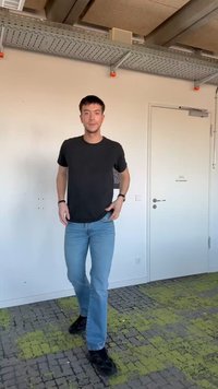 Bărbat tânăr care merge pe o mochetă verde și gri cu un model, purtând un tricou negru, blugi albaștri și adidași negri.