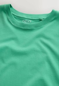 Vihreä puuvillainen t-paita pyöreällä kaula-aukolla, jossa näkyy sisäpuolen merkintä, johon on painettu tuotemerkin nimi "NEXT".