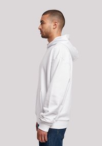 Young man with short hair wearing a white hoodie and blue jeans, standing in profile against a plain light gray background.