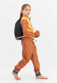 Lapsi, joka on pukeutunut oranssiin vedenpitävään takkiin ja siihen sopiviin housuihin mustilla yksityiskohdilla. Musta laukku on heitetty toisen olkapään yli.