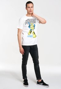 White cotton t-shirt featuring a yellow and blue graphic of a robot with text above, paired with dark jeans and black sneakers.