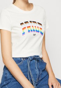 Bílý tričko s barevným grafikou "PRIDE" v duhových barvách a černým textem, doplněné o vysoké modré džíny s uvázaným pasem.