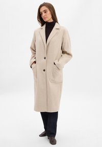 Beige Wollmischungmantel mit lockerer Passform, zwei großen Taschen, eingelegtem Revers und Ein-Knopf-Verschluss. Über einem schwarzen Rollkragenpullover getragen.
