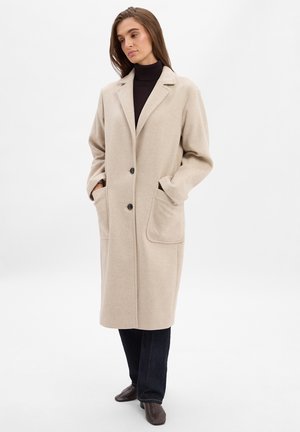 Beige Wollmischungmantel mit lockerer Passform, zwei großen Taschen, eingelegtem Revers und Ein-Knopf-Verschluss. Über einem schwarzen Rollkragenpullover getragen.