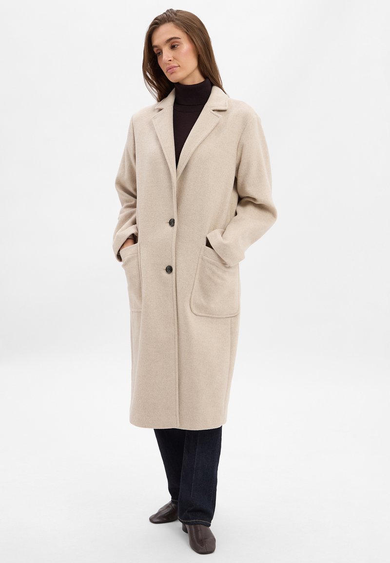 Beige Wollmischungmantel mit lockerer Passform, zwei großen Taschen, eingelegtem Revers und Ein-Knopf-Verschluss. Über einem schwarzen Rollkragenpullover getragen.