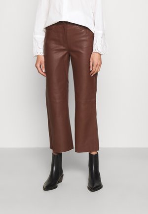 Femme portant un pantalon en cuir marron court, un chemisier blanc avec des poignets en dentelle et des bottines noires à talons, debout devant un fond uni.