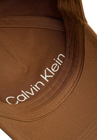 Casquette en tissu marron avec une visière courbée ; présente un texte "Calvin Klein" en blanc à l'intérieur. Les détails brodés améliorent le design global.