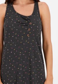 Kobieta w ciemnoszarej bezrękawnikowej bluzce z małymi wzorami liści w kolorze różowym, żółtym i szarym oraz trzema brązowymi guzikami z przodu.