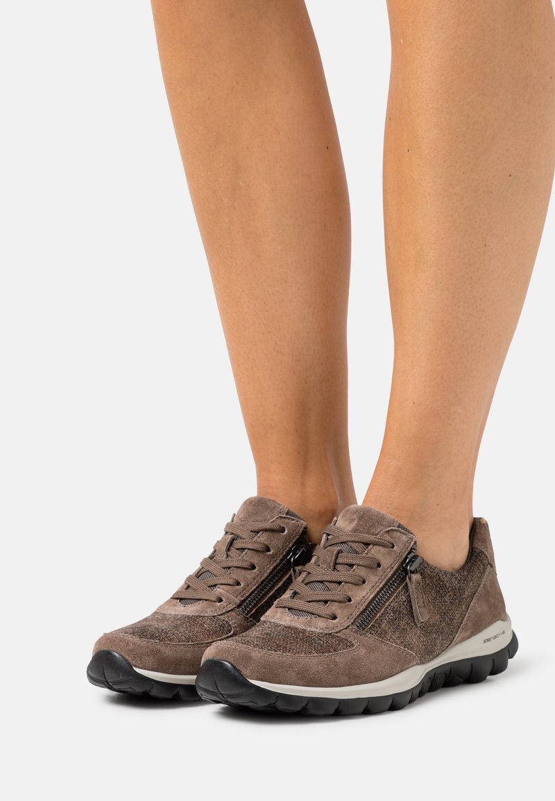 Baskets en daim marron avec semelles noires et fermetures éclair latérales, portées pieds nus sur fond blanc.
