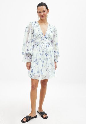 Robe à motifs floraux avec de longues manches transparentes, décolleté en V et taille froncée. Principalement blanche avec des fleurs bleues et violettes. Associée à des sandales noires.