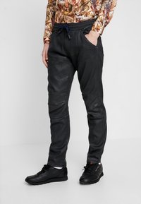 Los pantalones jogger de cuero negro presentan un ajuste ajustado, cintura elástica con cordón y paneles en las rodillas para un diseño adicional. Se llevan con zapatos negros.