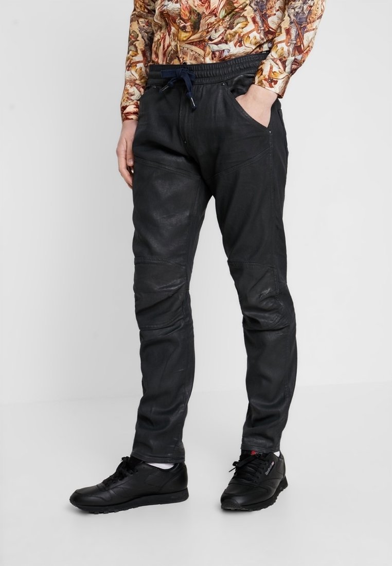 Los pantalones jogger de cuero negro presentan un ajuste ajustado, cintura elástica con cordón y paneles en las rodillas para un diseño adicional. Se llevan con zapatos negros.