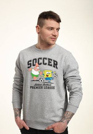Ung mand iført en grå sweatshirt med SpongeBob og Patrick, der spiller fodbold, og teksten "Soccer Bikini Bottom Premier League."