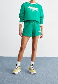 Osoba ubrana w zieloną bluzę Nike i pasujące zielone szorty, białe skarpetki oraz żółto-białe sneakersy stoi na niebieskim dywanie.