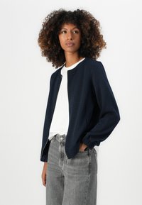Frau mit lockigem Haar, die eine marineblaue Zip-Jacke über einem weißen Hemd und grauen, hoch taillierten Jeans trägt, steht mit einer Hand in der Tasche.