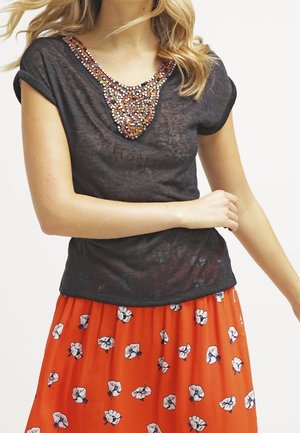 Czarna bluzka z krótkim rękawem z wielokolorowymi koralikami wokół dekoltu, zestawiona z jasnopomarańczową spódnicą w białe kwiatowe wzory.
