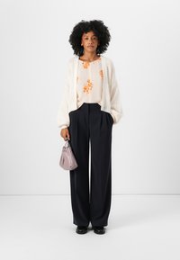 Cardigan alb tricotat peste o bluză florală piersicie, asociată cu pantaloni navy cu talie înaltă și o geantă lila. Pantofi negri completează ținuta.