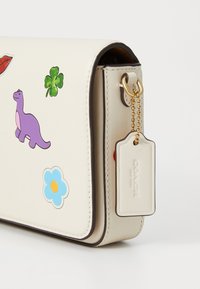 Beige leather crossbody bag with colorful stickers including a dinosaur, flower, and lips; gold-tone hardware with a branded tag.