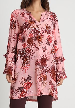 Różowa tunika z czerwonymi i brązowymi kwiatowymi wzorami, długie rozszerzane rękawy z falbankami, noszona na ciemnych spodniach.