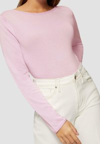Jasnoróżowa bluzka z długim rękawem w dopasowanym kroju, wykonana z gładkiego, elastycznego materiału. Połączona z białymi spodniami z wysokim stanem.