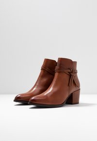 Pari lakkautettuja ruskeita nahkasaappaita nilkkakorkeudella, joissa on tukevat korot ja kietoutuva nauhadesign, esillä valkoisella pinnalla.