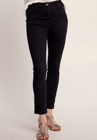 Jeans skinny noirs taille haute avec une finition lisse, cinq poches et une fermeture à bouton à l'avant, associés à des talons métalliques à brides.