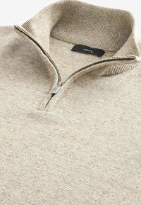Beige zip-hals pullover med ribbet krave og kant. Blød tekstur, subtil sprøjtet mønster og metal lynlås detalje. Brand mærke synligt.