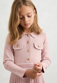 Rochie roz cu mâneci lungi, guler, închidere cu nasturi aurii, buzunare la piept și detalii de cusături albe; fustă plisată.