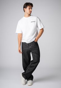 Weißes T-Shirt mit kurzen Ärmeln und dem Text "DOWNTOWN CLUB", kombiniert mit lockeren schwarzen Jeans und weißen Sneakers, vor einem neutralen Hintergrund.