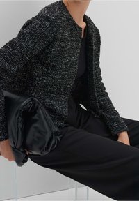 Veste texturée noire et blanche à encolure ronde, associée à un haut et un pantalon noirs, tenant une pochette en tissu lisse noir.