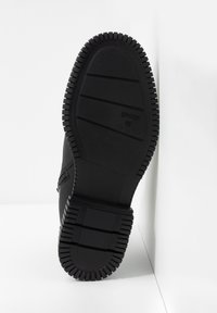 Suela de zapato negra con profundos surcos, con la talla 42 marcada, posicionada en vertical contra un fondo blanco en la esquina.