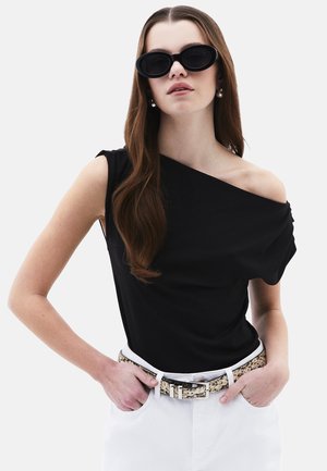 Femme aux longs cheveux bruns portant des lunettes de soleil noires à monture œil de chat, un haut noir à une épaule, des boucles d'oreilles en perles, un pantalon blanc et une ceinture à paillettes.